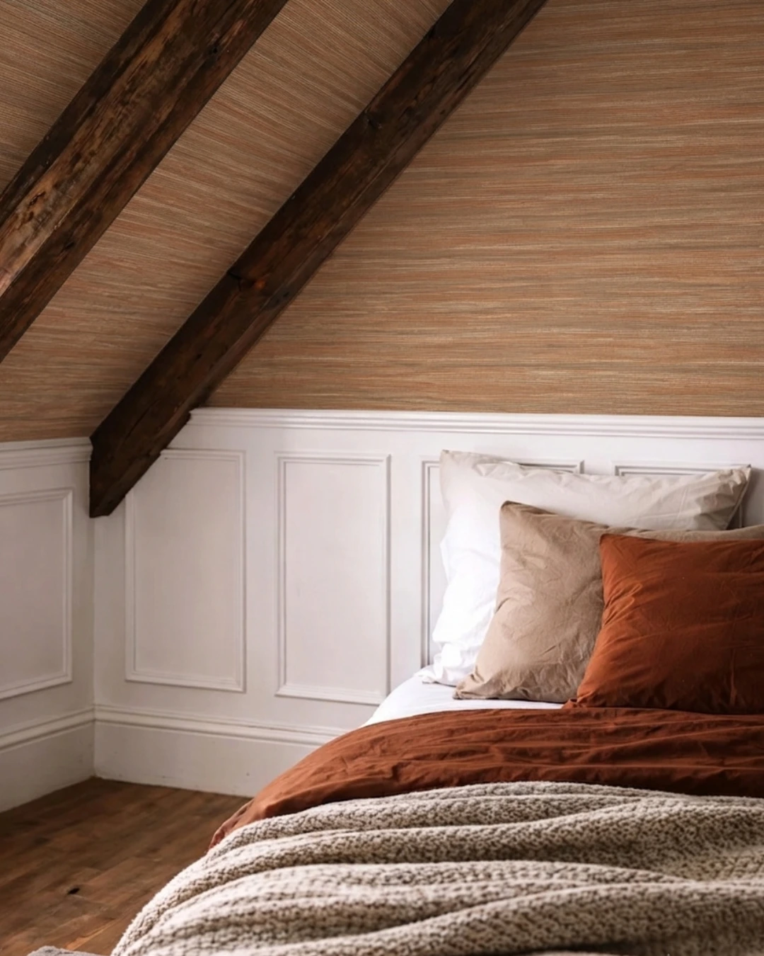 Attic bedroom with sloped ceiling lined in beige-orange grasscloth and dark wooden beams