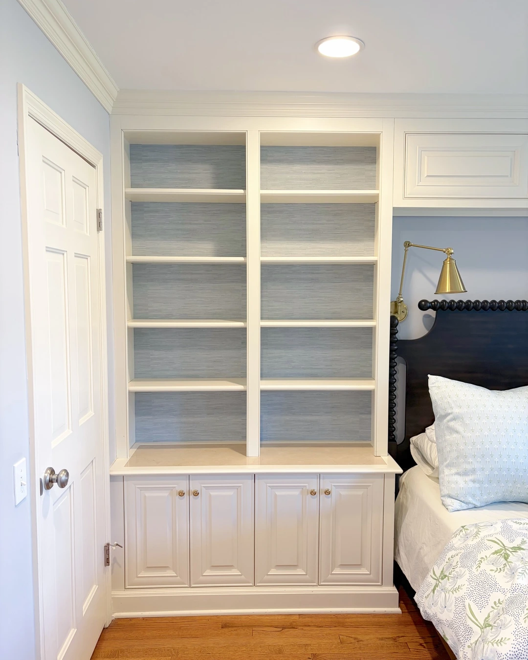White built-in bookshelves backed in light blue-gray grasscloth beside a bed