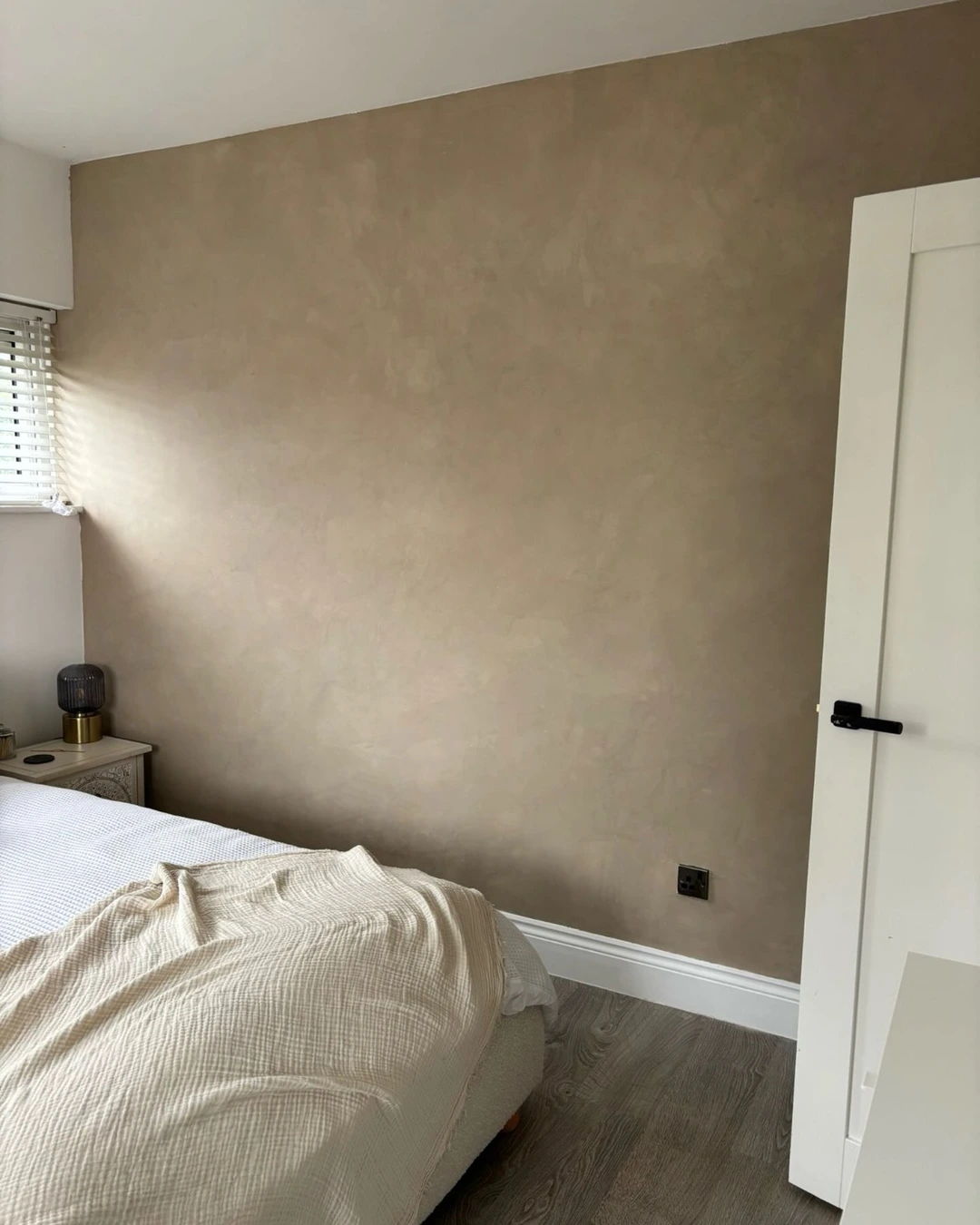 Bedroom with a smooth warm taupe microcement feature wall behind a bed with white bedding