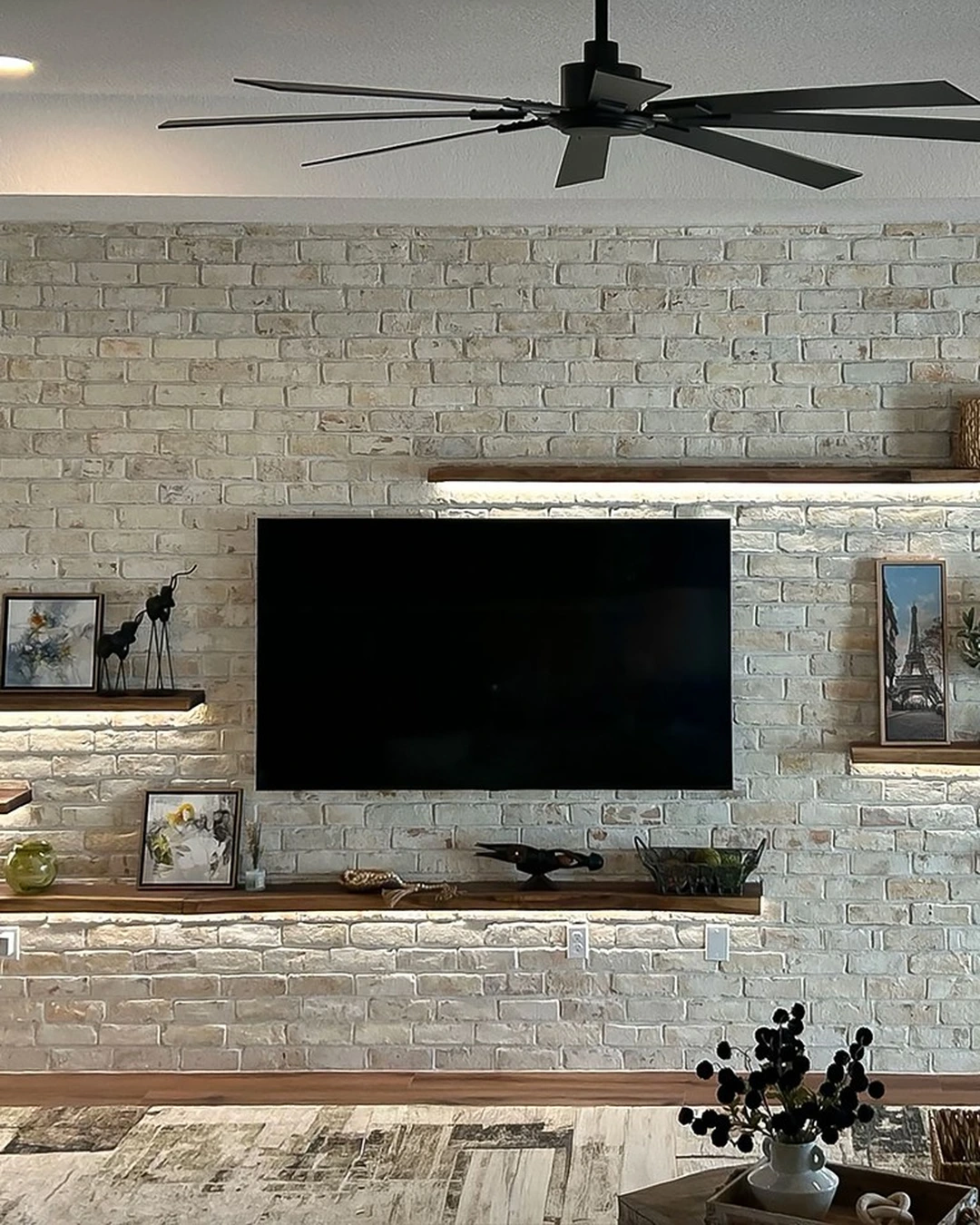 Whitewashed brick feature wall with a mounted television, lit floating wood shelves and decor