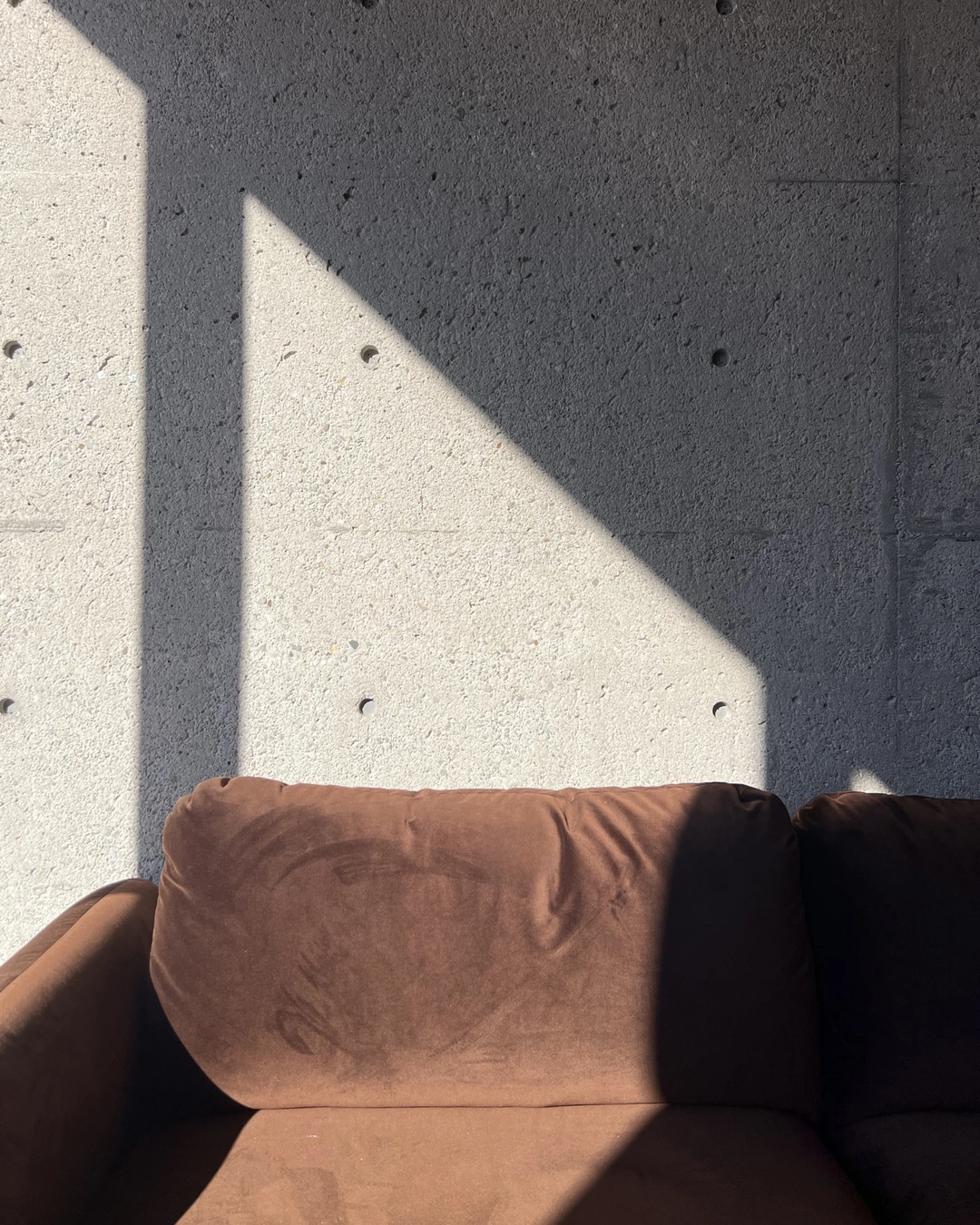 Raw board-formed concrete wall with pitted surface and tie-hole grid above a brown velvet sofa