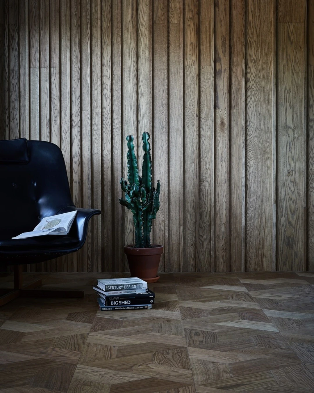 Dark vertical wood slat wall behind a black leather lounge chair, tall cactus and stacked books