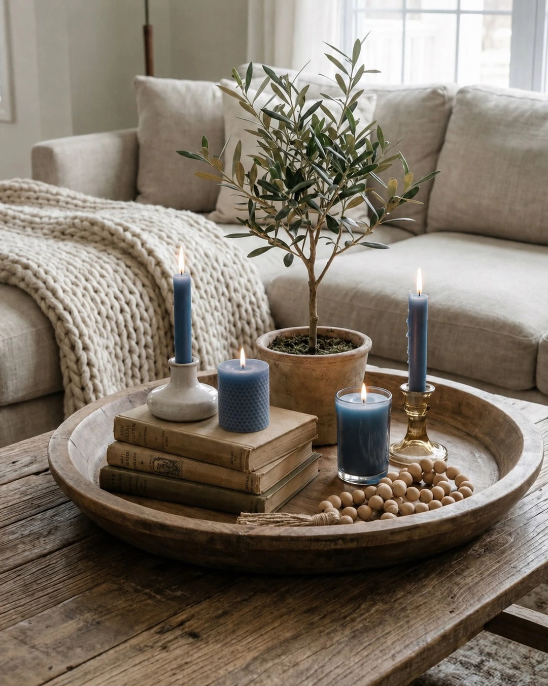 Round wooden tray with four lit blue candles in different holders, stacked books, wooden beads, and a potted olive tree