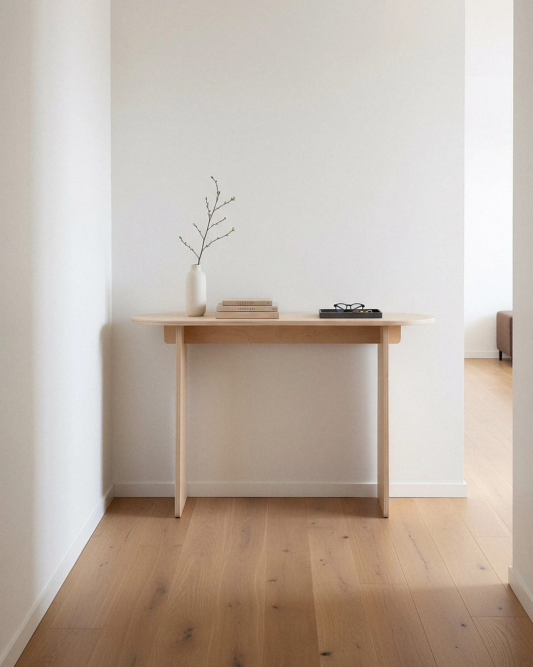 Light wood console against a white wall with a white vase holding a bare branch, a stack of books, and a small black tray