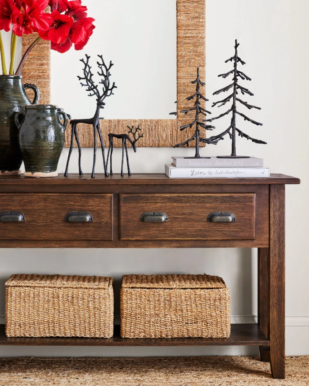 Dark wood console under a rattan-framed mirror with green glazed vases, black metal deer and tree sculptures on books, and woven baskets below