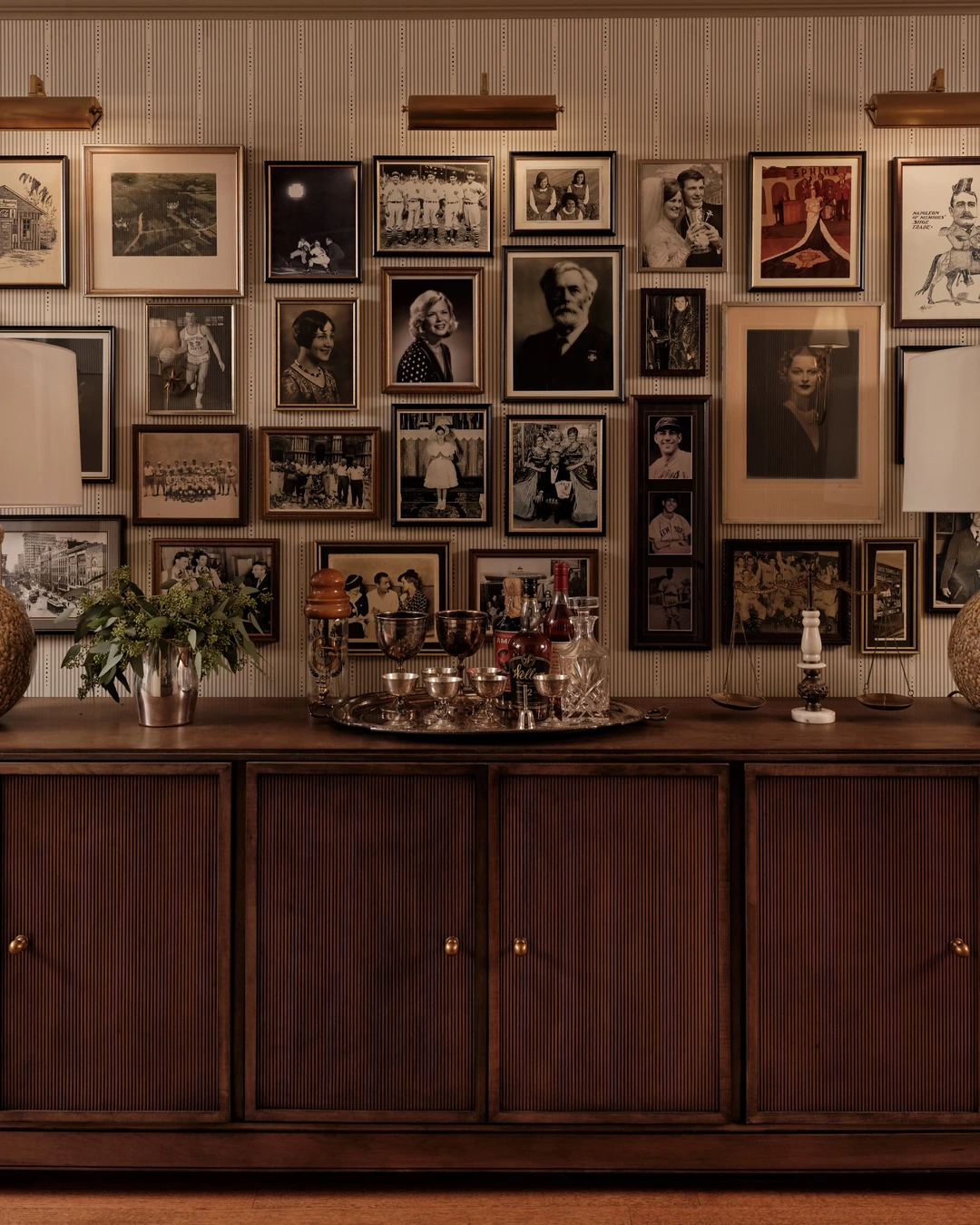 Dense gallery wall of framed black-and-white photographs and portraits above a dark wood sideboard with lamps