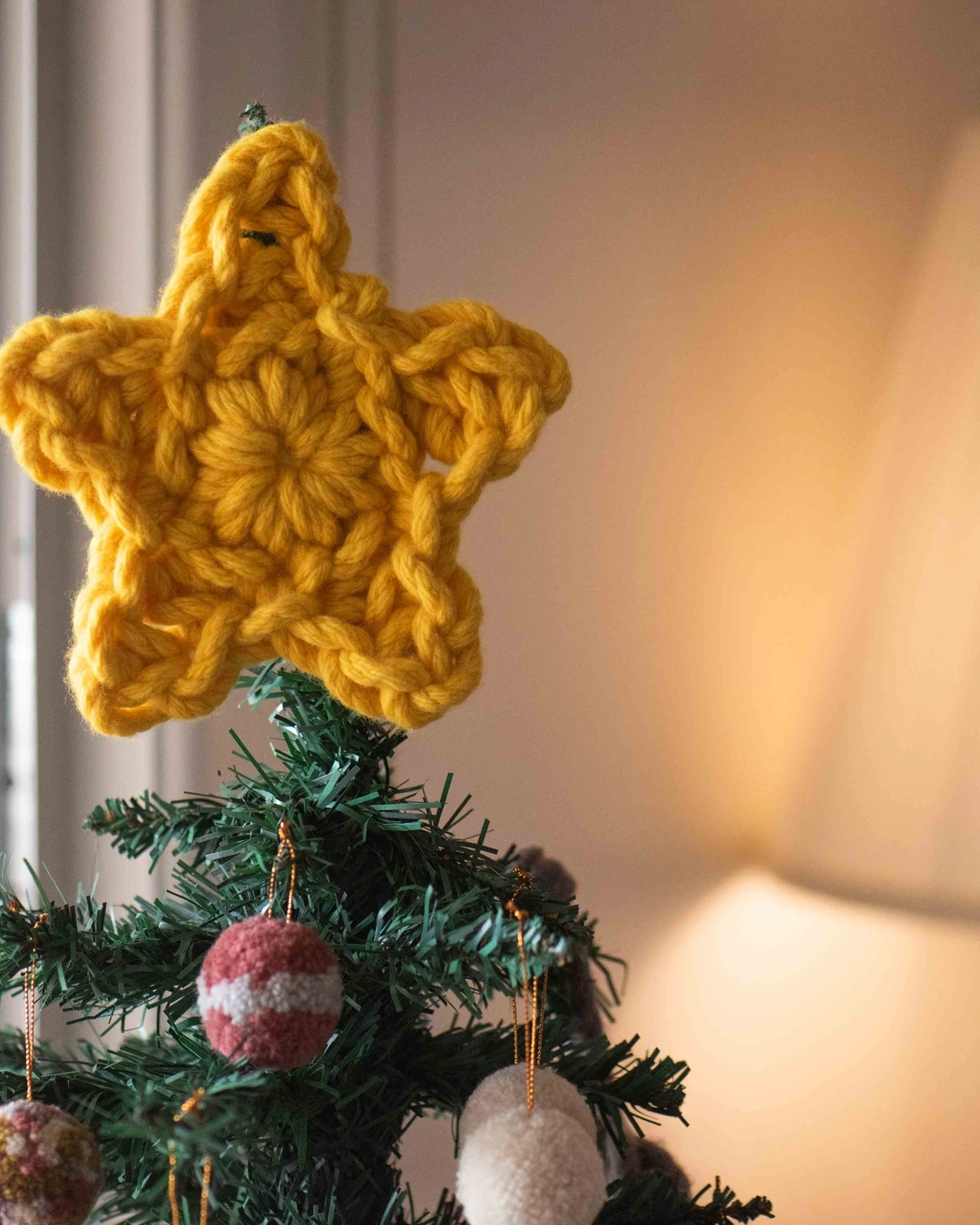 Chunky mustard-yellow crocheted star on top of a small artificial Christmas tree with pom-pom ornaments