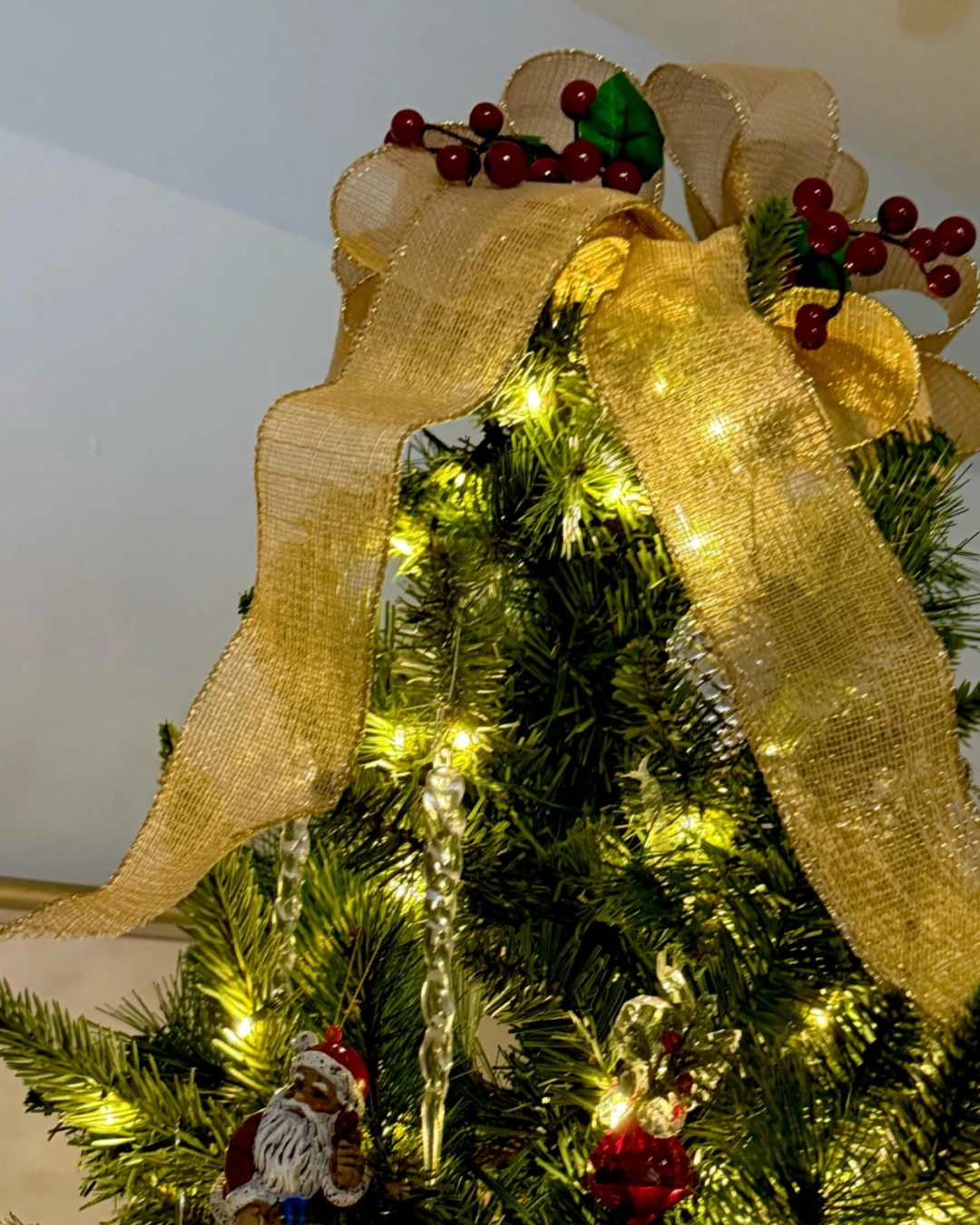 Gold burlap bow with holly and red berries crowning a lit Christmas tree, Santa and icicle ornaments below