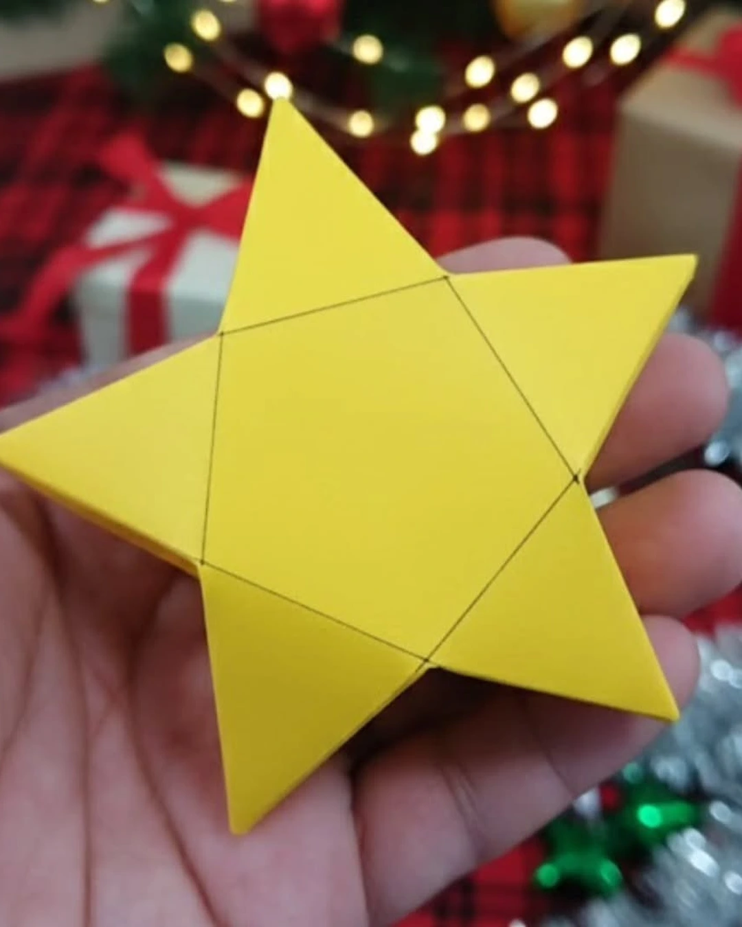 Bright yellow origami paper star held in a hand with wrapped gifts and fairy lights behind
