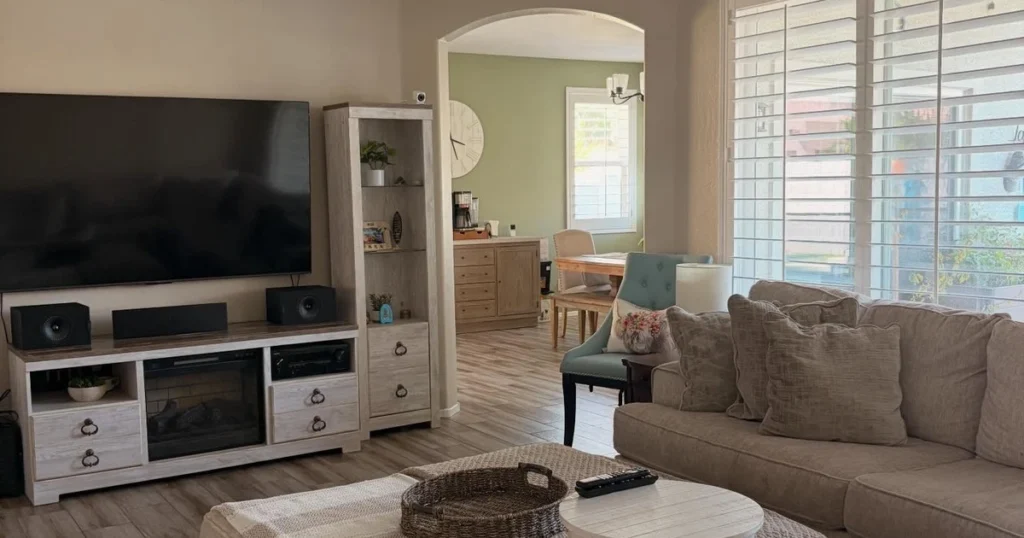 Beige living room with a wall-mounted TV, light wood console, and sofa, with a green dining area visible through an arched opening.