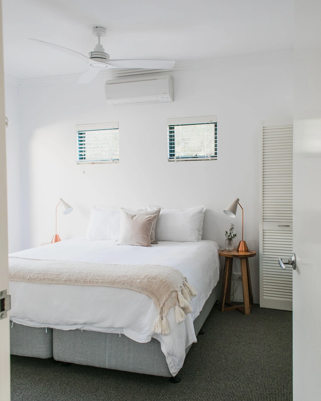 Bright white bedroom with white bedding, a beige throw, copper-toned lamps, and a wall air-conditioning unit.