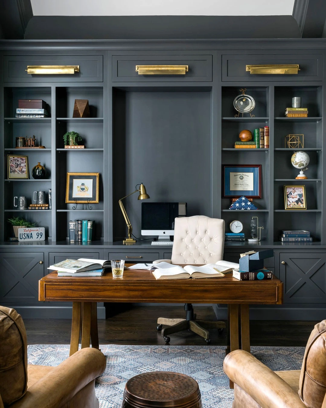 Home office with dark bronze-gray built-in bookcases, a wooden desk, a tufted office chair, and leather armchairs.