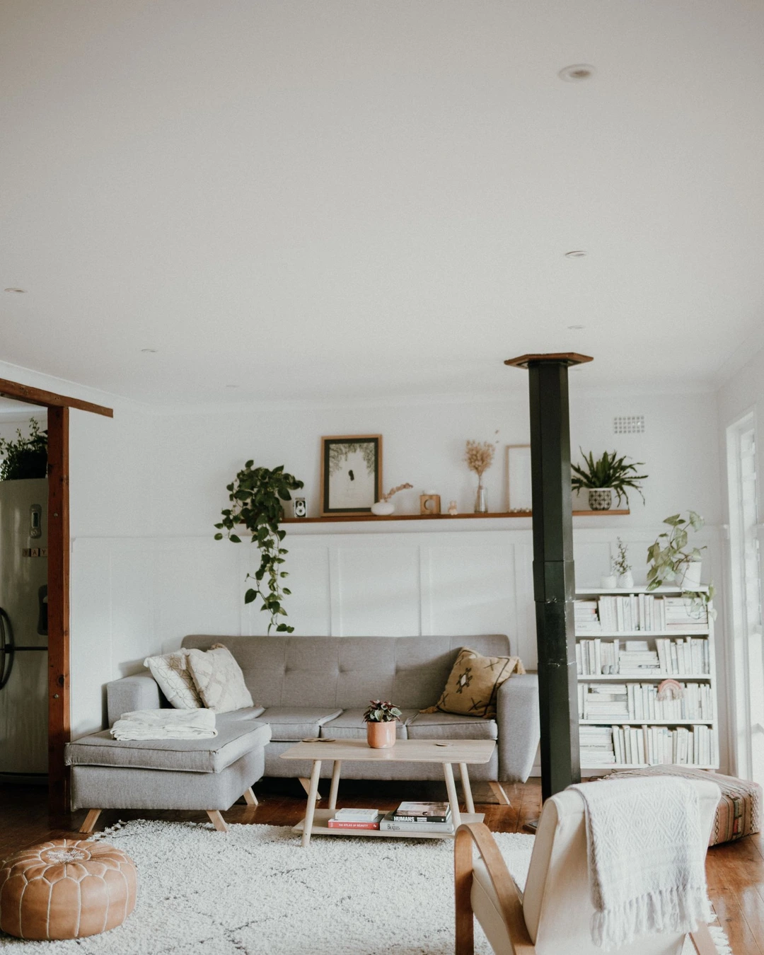 Warm off-white Scandinavian living room with a gray sofa, plants, and a white bookshelf, with a black stove pipe in the center.