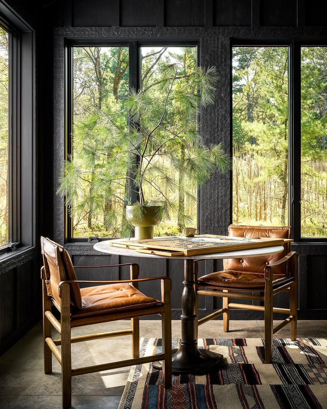 Sunroom with black paneled walls, large windows over green trees, brown leather armchairs, and a round marble-top table.