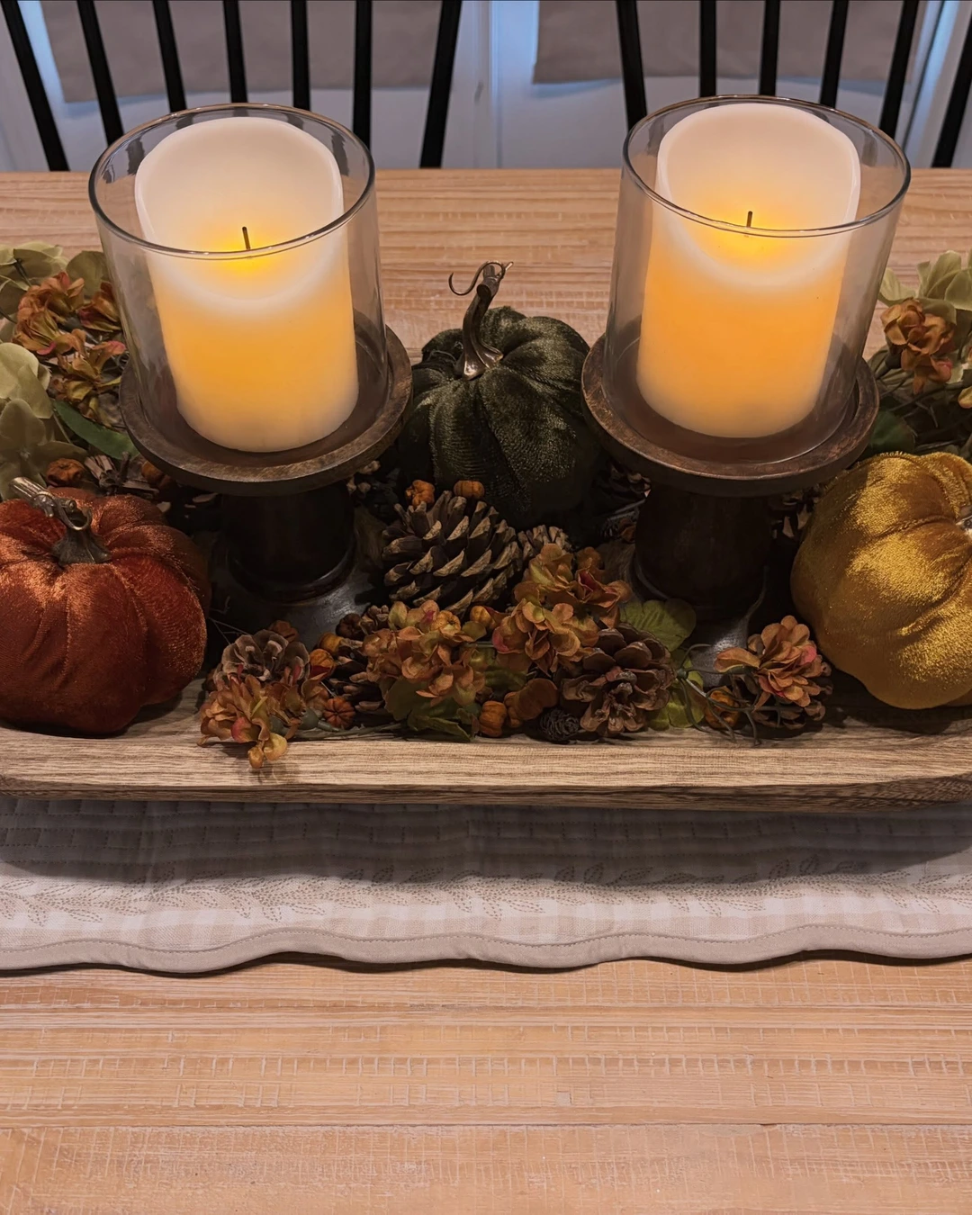 Light wood dough bowl with two LED pillar candles in glass holders, rust, gold, and green velvet pumpkins, pinecones, and dried flowers.