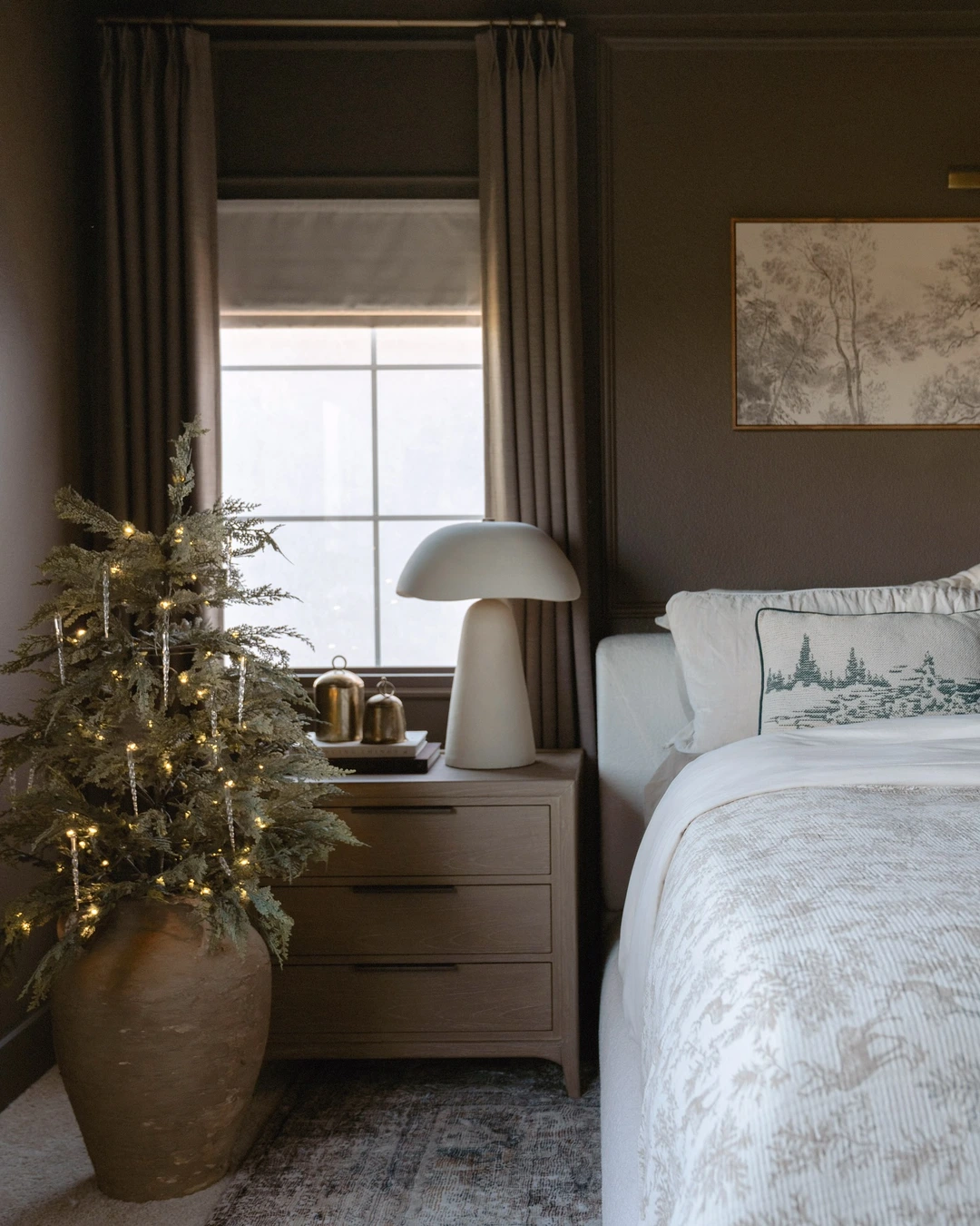 Small flocked tree in terracotta vase on floor beside wooden nightstand with lamp, books, and brass bells, beside a bed.