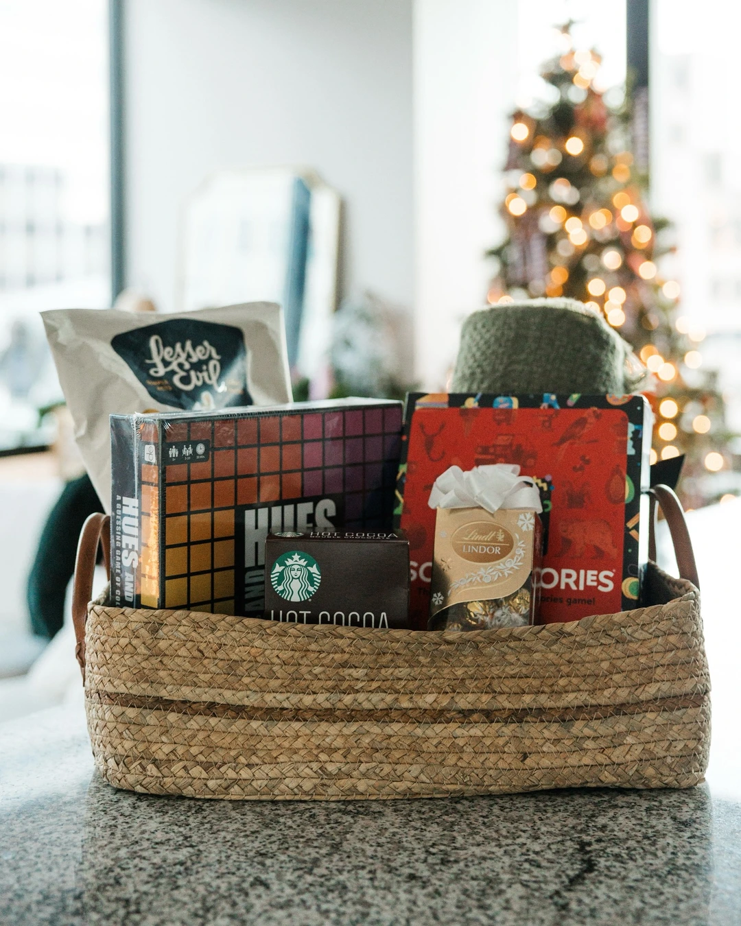 Woven basket with board games, popcorn, hot cocoa, truffles, and a green throw, with a lit Christmas tree behind it.