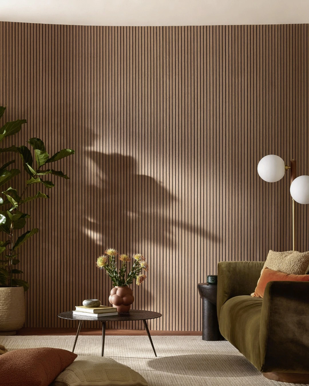 Vertical wood slat accent wall behind an olive-green sofa, a round coffee table with books and flowers, and plant shadows