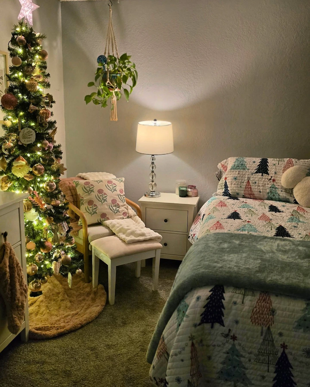 Armchair with floral pillow and throw beside nightstand with lit lamp, between lit Christmas tree and bed with Christmas-tree quilt.