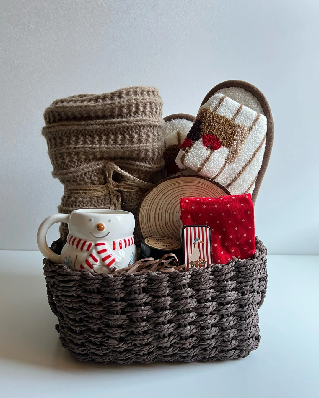 Dark woven basket with a folded knit blanket, fuzzy slippers, a snowman mug, red socks, a wood-slice coaster, and a candle tin.