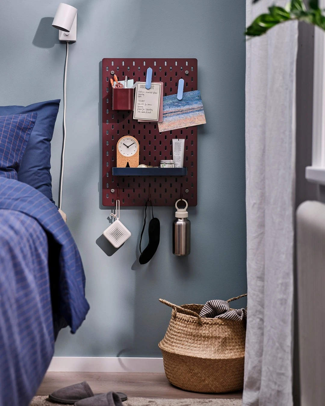Burgundy pegboard beside a bed holding a clock, clipped notes, toiletries, a speaker, a sleep mask, and a water bottle