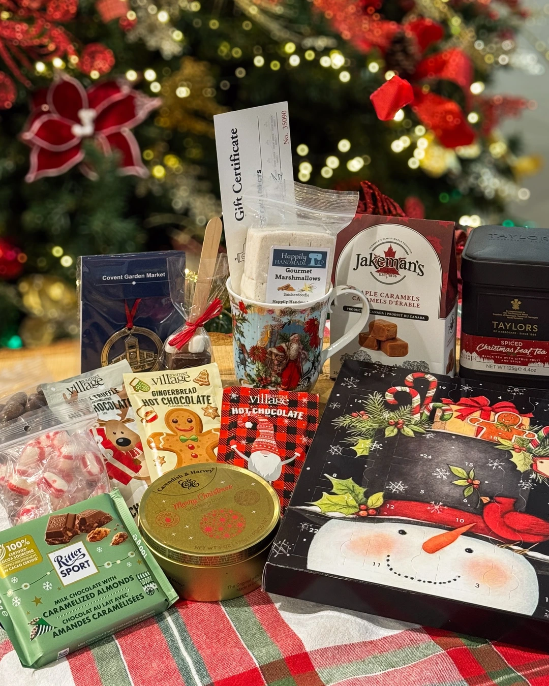 Sweet treats laid out on a plaid cloth in front of a lit tree, including a Santa mug with marshmallows, caramels, tea, chocolate, and an advent calendar. No basket is visible in this image.