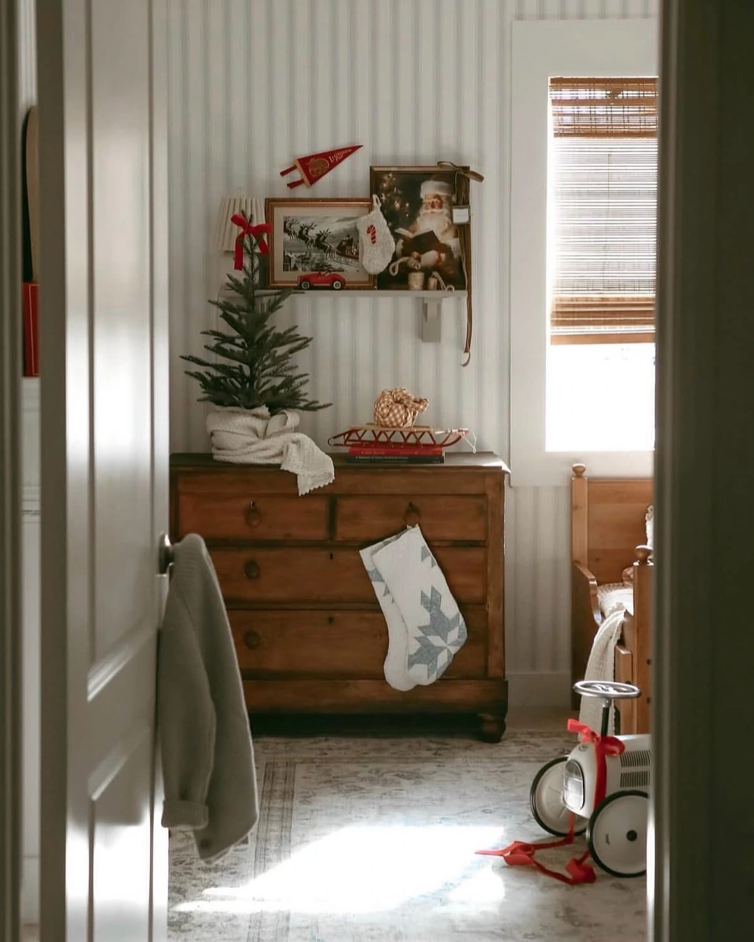 Wooden dresser with small tree in white cloth, red sled, books, and one white stocking with grey snowflake on a drawer pull.
