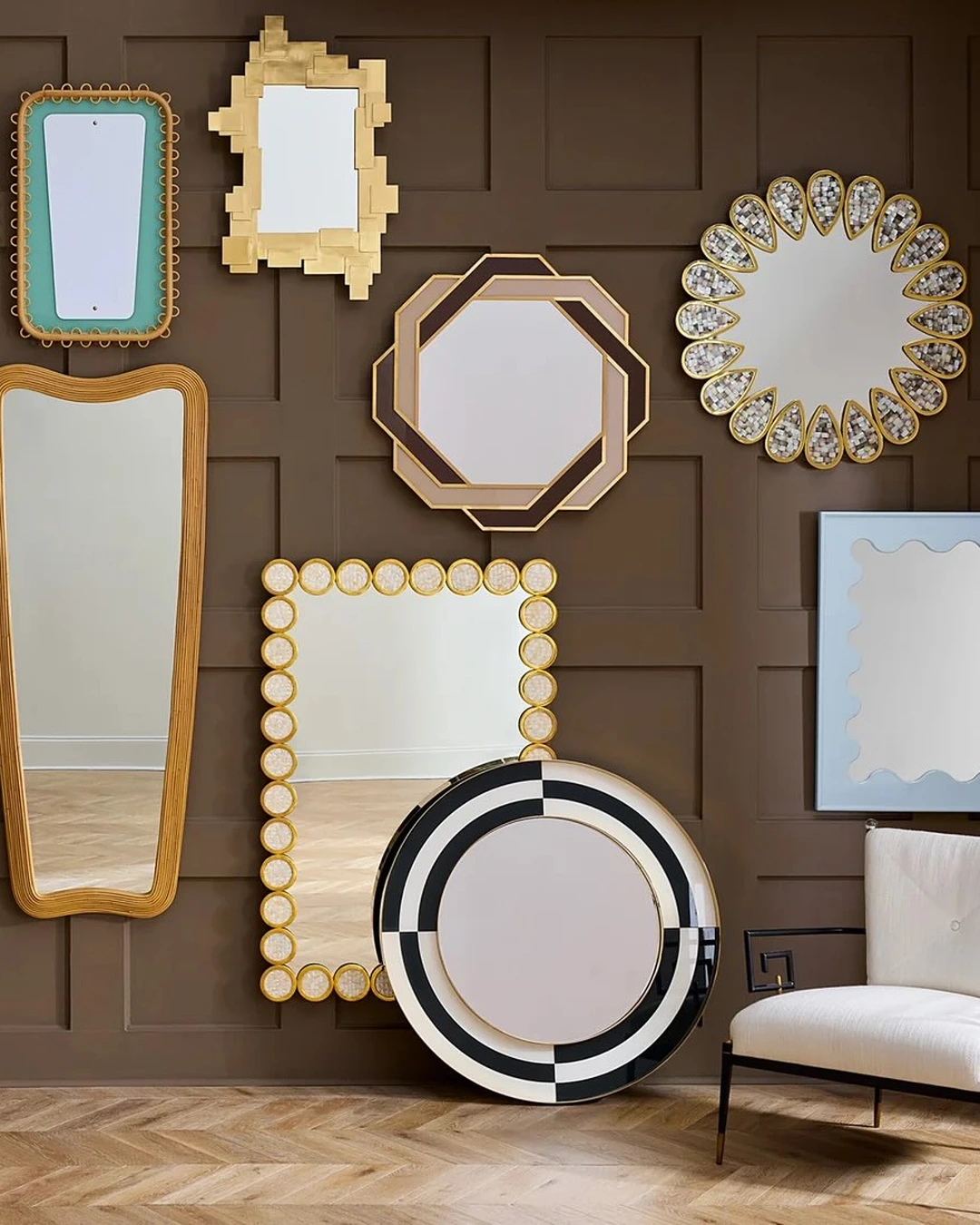 Gallery of decorative mirrors in round, octagonal, and wavy frames on a dark brown paneled wall, with a white chair below and a large round mirror leaning against the wall