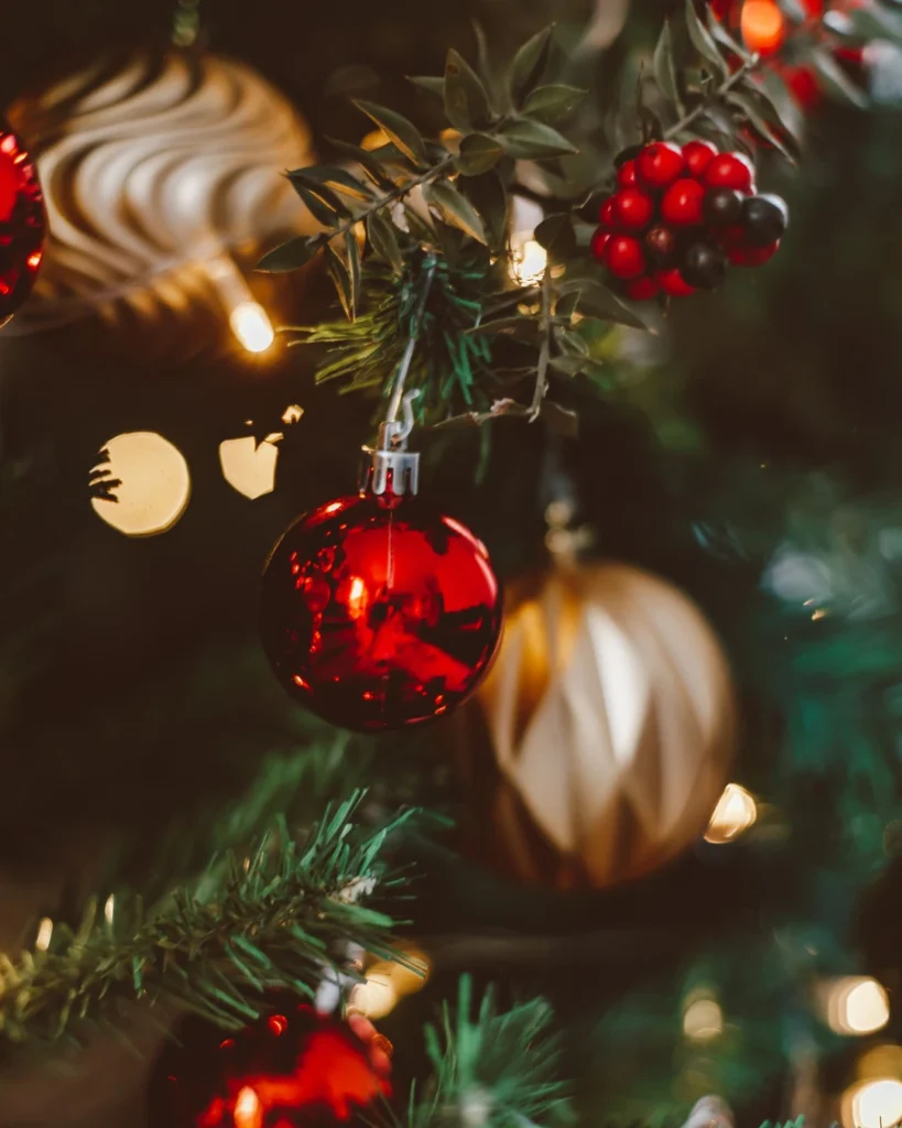 Close-up of a Christmas tree with shiny red and gold baubles, red berries, and warm lights among green branches.