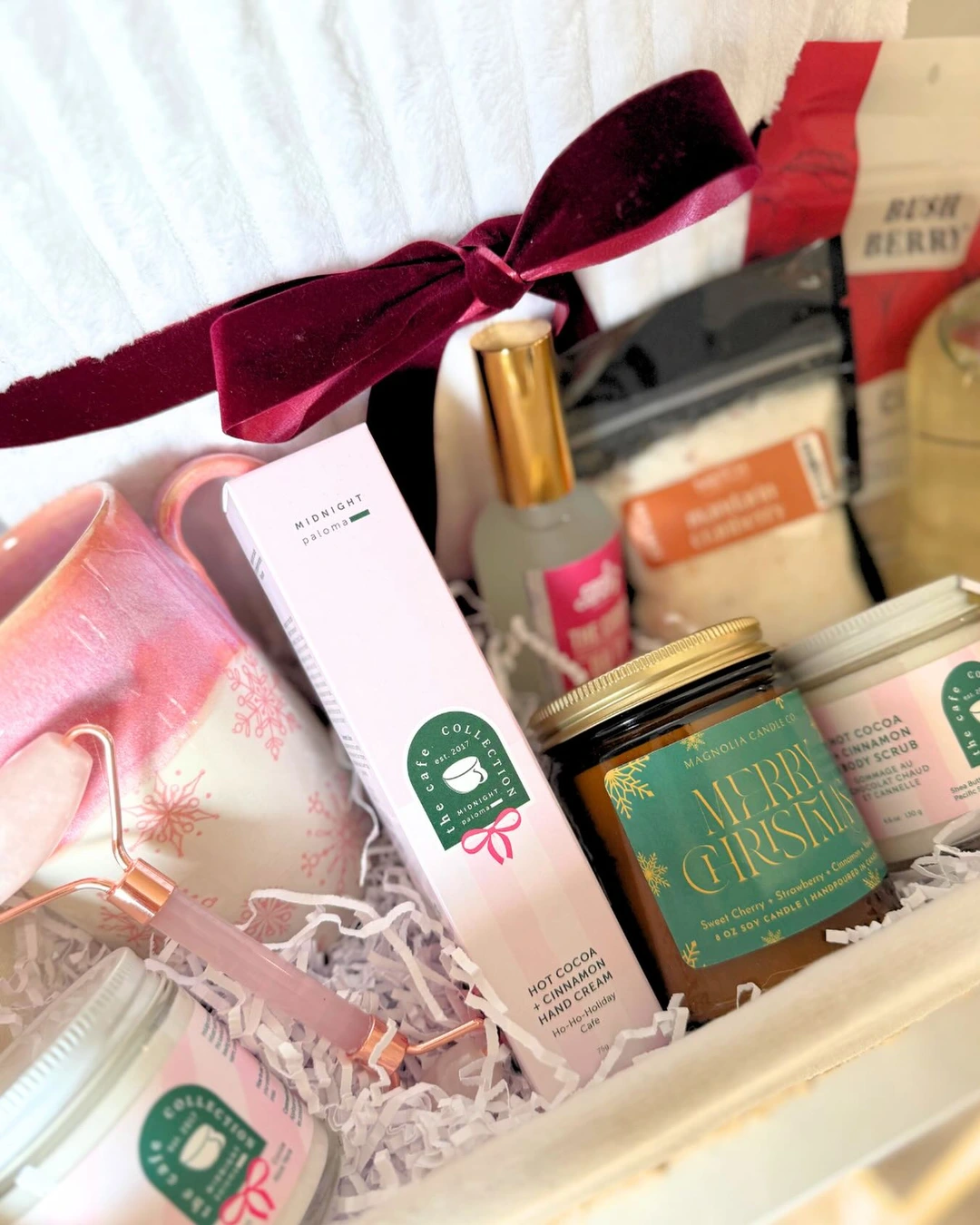 Spa basket with a pink snowflake mug, facial roller, hand cream, body scrub, and a white towel tied with a burgundy ribbon.