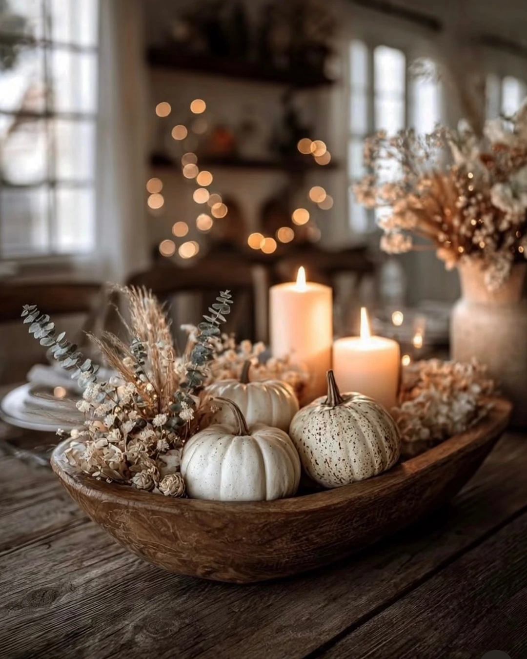 Carved wooden dough bowl with white speckled pumpkins, dried eucalyptus, wheat, and cream flowers, with lit candles behind it.