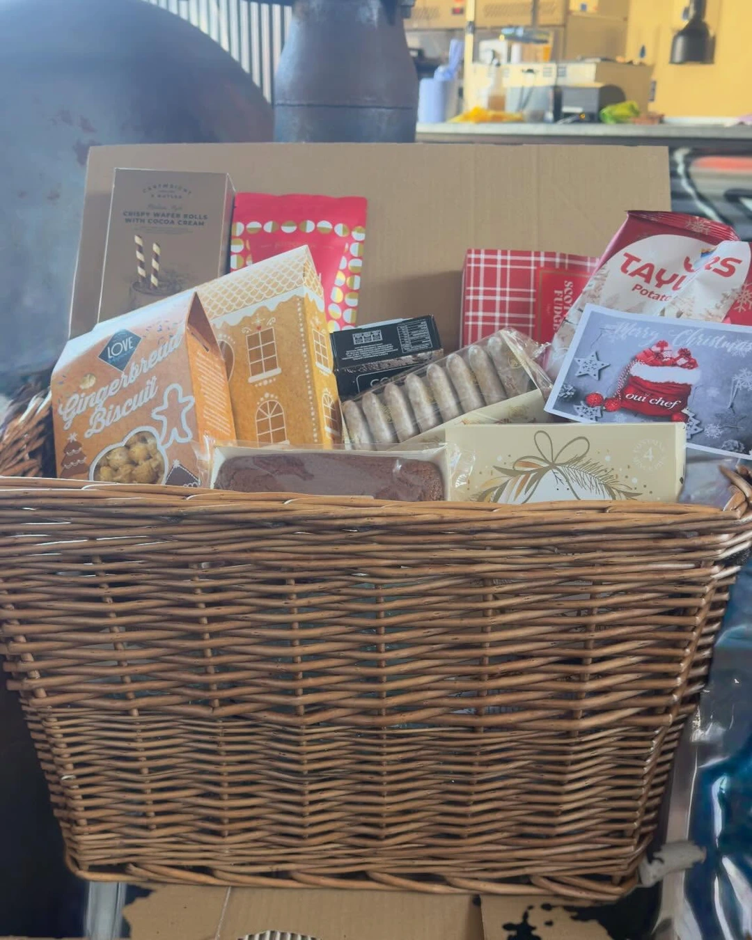 Large wicker basket overflowing with gingerbread biscuits, a gingerbread-house box, wafer rolls, fudge, biscuits, and a cake loaf.