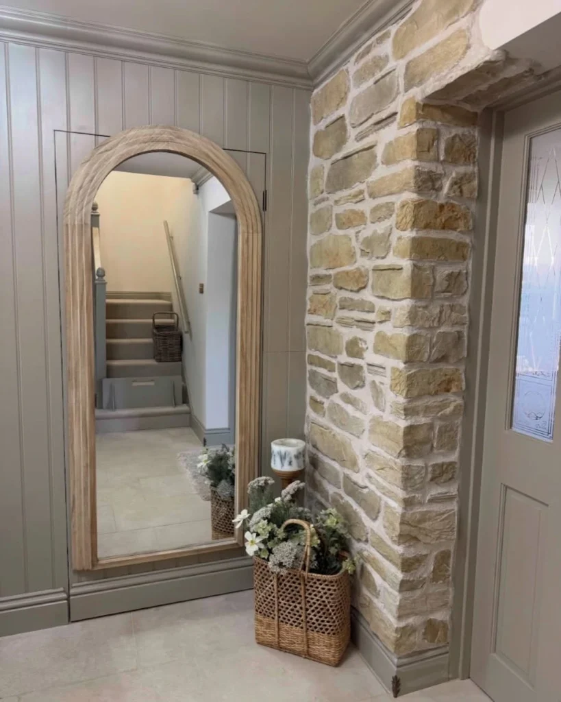 Large arched floor mirror with a light wood frame on a gray paneled hallway wall, reflecting a staircase and a wicker basket
