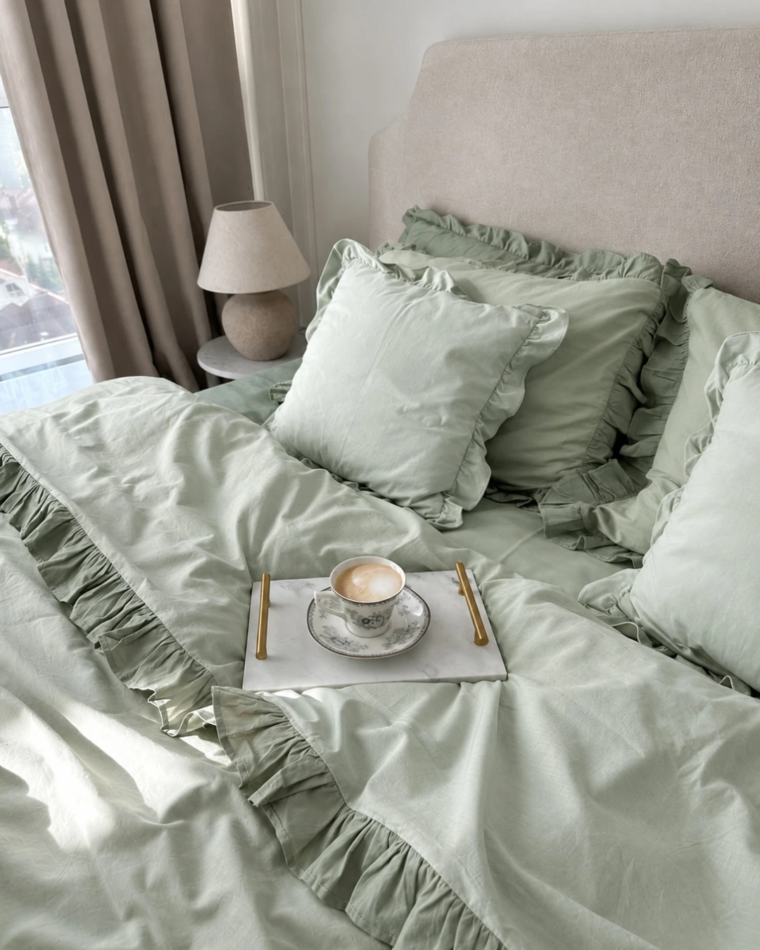 Sage-green linen bedding with ruffle trim on the duvet and pillows beside a bedside lamp and window