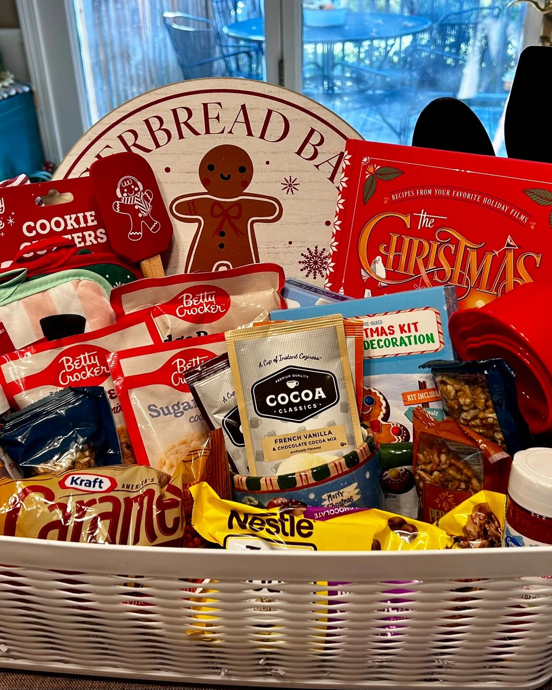White woven basket overflowing with baking mixes, cookie cutters, a red spatula, oven mitts, measuring cups, a whisk, and sprinkles.