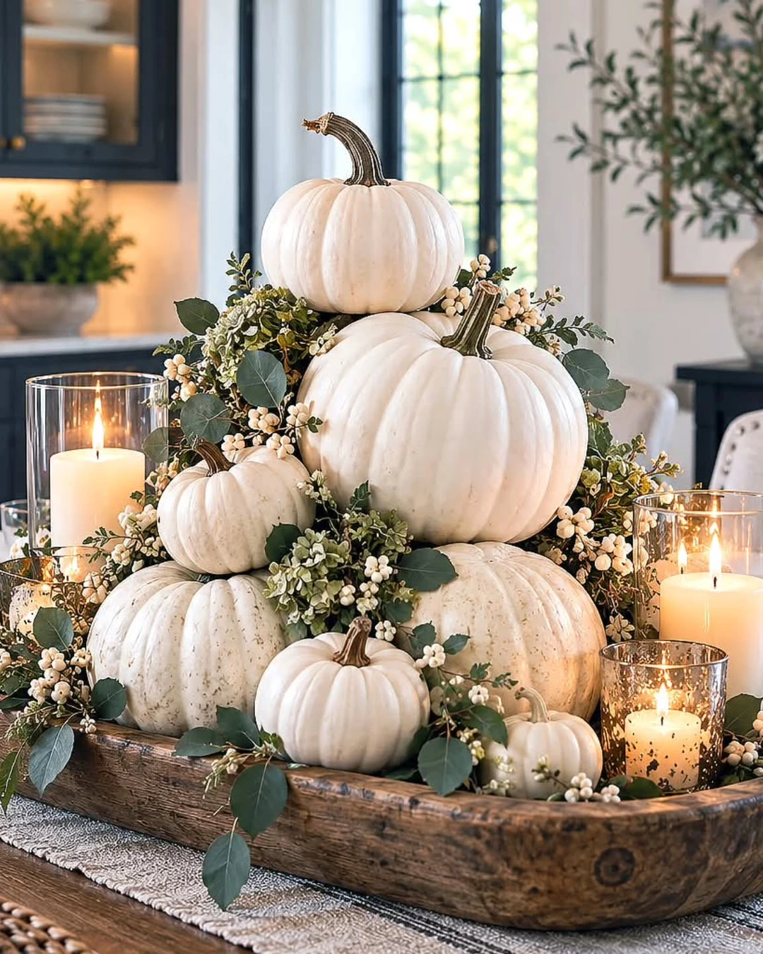 Wooden dough bowl stacked with white pumpkins of different sizes, green foliage, eucalyptus, white berries, and lit pillar candles in glass holders.