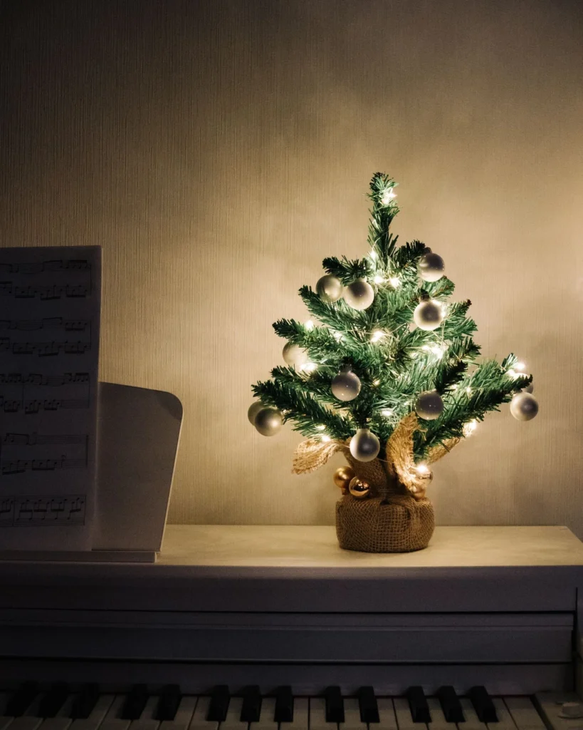 Small Christmas tree with white baubles and warm lights in a burlap-wrapped pot on a piano.