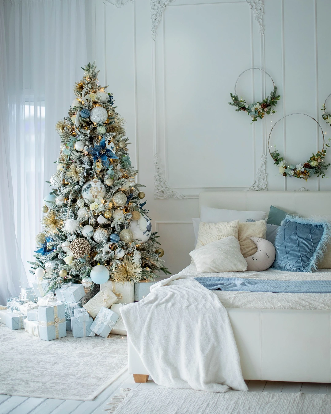 White bedroom with blue pillows and throw, tall tree with blue, gold, and white ornaments, gifts, and hoop wreaths.