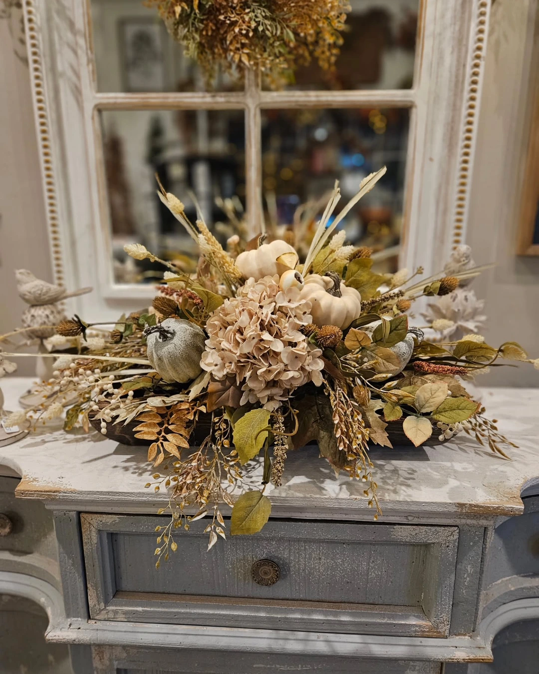 Dark wooden dough bowl on a dresser with cream pumpkins, large pale hydrangeas, dried wheat stalks, berries, and mixed foliage.