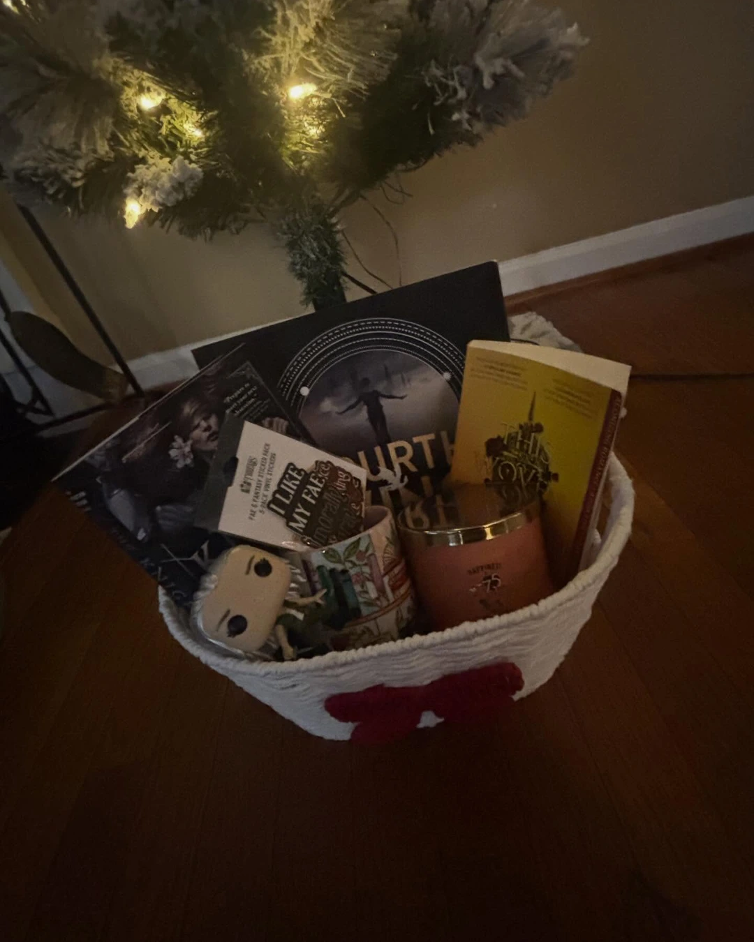 White rope basket with a red bow under a lit flocked tree, filled with fantasy books, a bookshelf-print mug, a peach candle, and a figurine.
