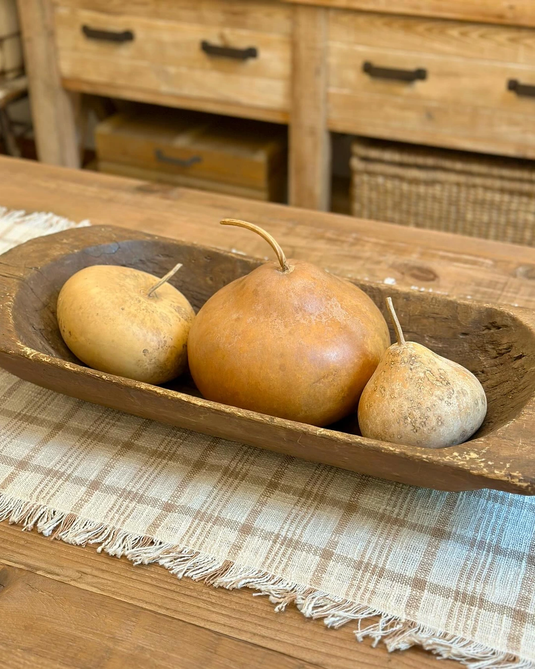 Weathered wooden dough bowl with three large tan and brown gourds on a beige and brown plaid fringed runner.