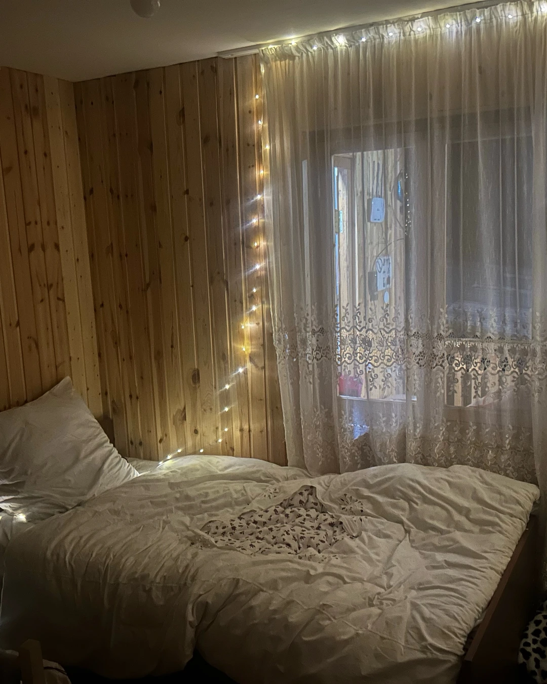 Small bedroom with bed beside window covered in sheer lace curtains with warm fairy lights, wood panel walls.